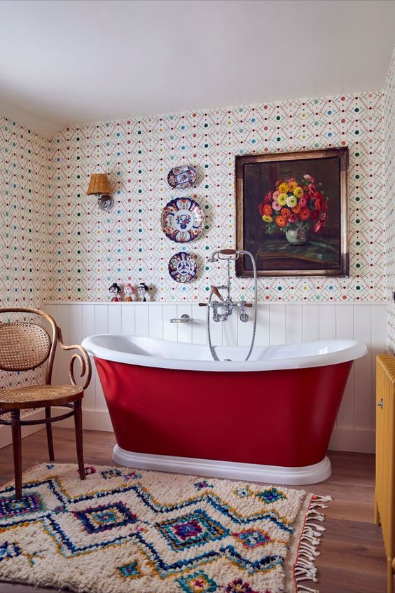 a sophisticated vintage bathroom with bold wallpaper and white shiplap, a burgundy bathtub, a colorful rug and plates and an artwork