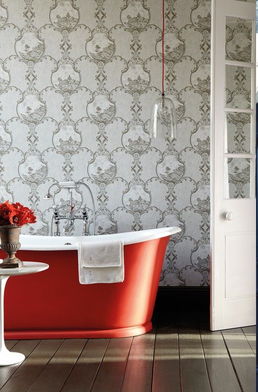 a refined bathroom with grey printed wallpaper, a stained floor, a red free-standing bathtub, a glass pendant lamp over the tub
