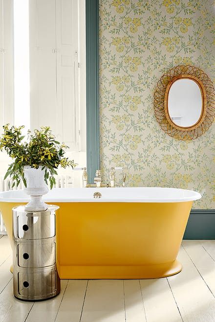 an elegant vintage bathroom with floral wallpaper, a mirror in a woven frame, a mustard free-standing bathtub, a shiny side table and greenery