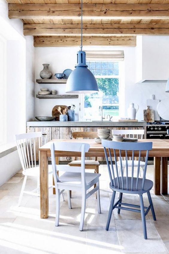 a beautiful coastal kitchen with modern concrete and wood cabinets, a rustic dining table, mismatching vintage chairs and a blue lamp