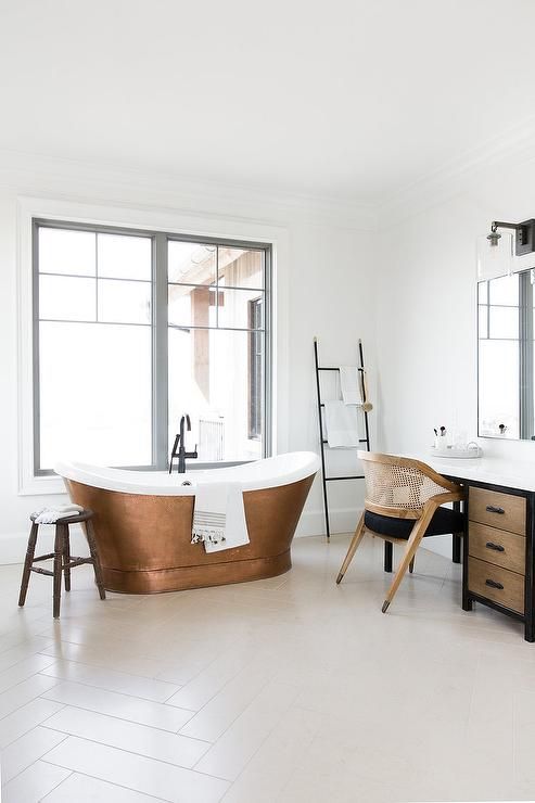 a beautiful light-filled bathroom in neutrals, with a wooden vanity and a stained and black chair, a cool copper bathtub and a ladder