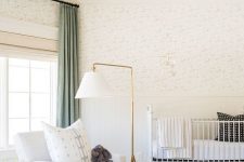 a beautiful neutral mid-century modern nursery with printed wallpaper, a white crib, a creamy chair, side tables, a gold floor lamp and a wicker pendant lamp
