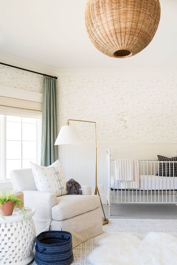 a beautiful neutral mid century modern nursery with printed wallpaper, a white crib, a creamy chair, side tables, a gold floor lamp and a wicker pendant lamp