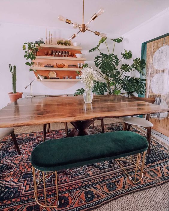 a boho mid-century modern dining room with a round orange shelving unit, a stained table, stained chairs and a green upholstered bench plus various plants