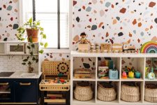 a bright mid-century modern nursery with a terrazzo accent wall, a white shelving unit, a navy play kitchen, some greenery and bright toys