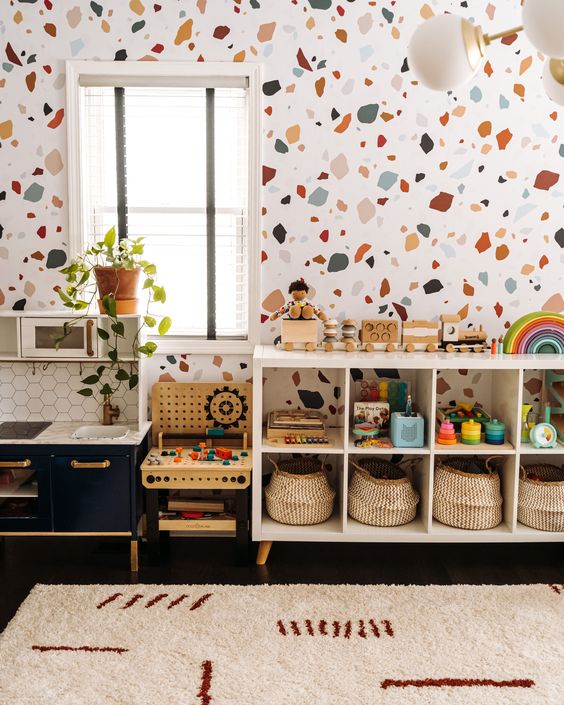 a bright mid-century modern nursery with a terrazzo accent wall, a white shelving unit, a navy play kitchen, some greenery and bright toys