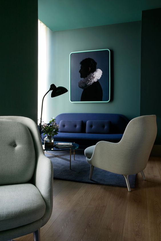 a chic and stylish living room with green walls, a navy loveseat, grey chairs, a glass coffee table and a bold and contrasting artwork