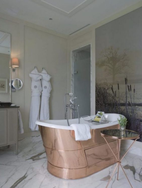 a delicate and neutral bathroom with tan walls and a gorgeous artwork on one of them, a shiny copper bathtub, a neutral vanity with a mirror