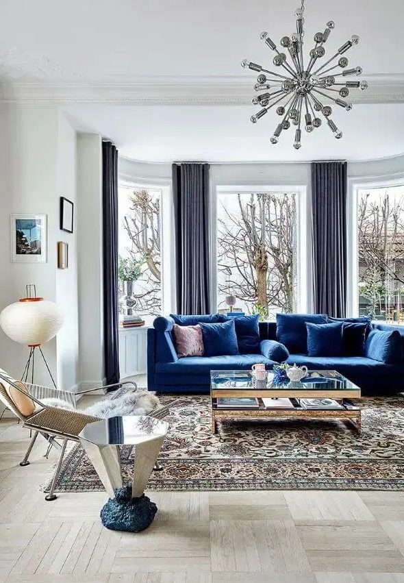 a light-filled living room wiht a bay window, a bold blue sofa, a glass coffee table and a mirror side table, a cool chair and a mid-century modern printed rug
