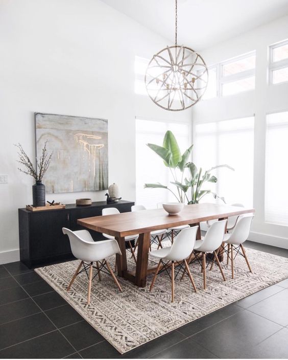 a mid-century modern dining space with a stained table and white Eames chairs, a printed rug, a black credenza, a sphere chandelier and potted plants
