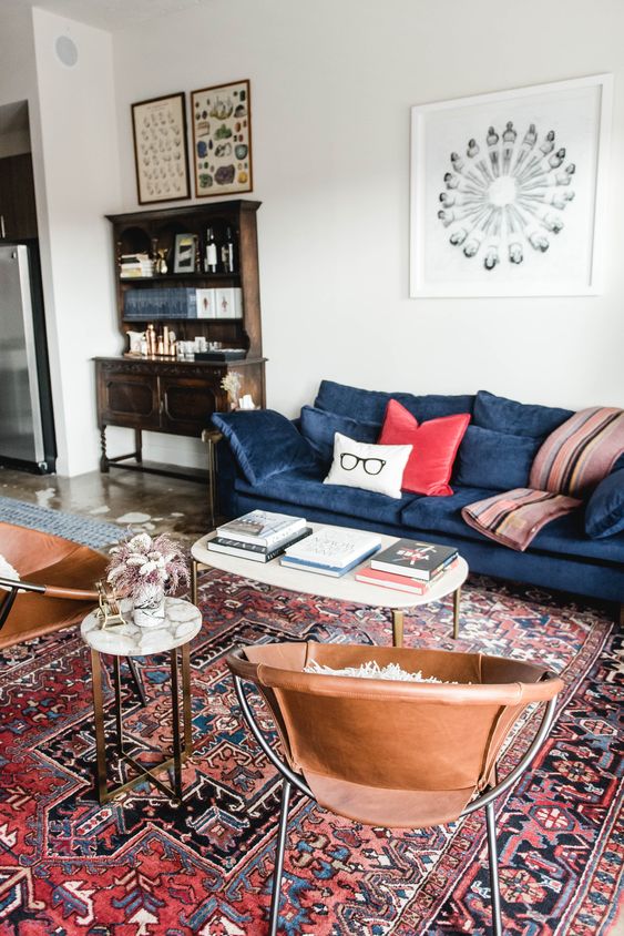 a mid-century modern living room with a vintage bureau, a blue sofa with colorful pillows, leather chairs, coffee tables and a red printed rug
