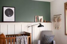 a mid-century modern meets boho nursery with a green and paneled accent wall, a stained crib, a grey chair, a leather Moroccan pouf and a black floor lamp