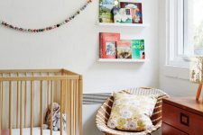 a mid-century modern nursery with open shelves for books, a stained crib, a stained dresser, a wicker round chair, printed textiles and lovely toys