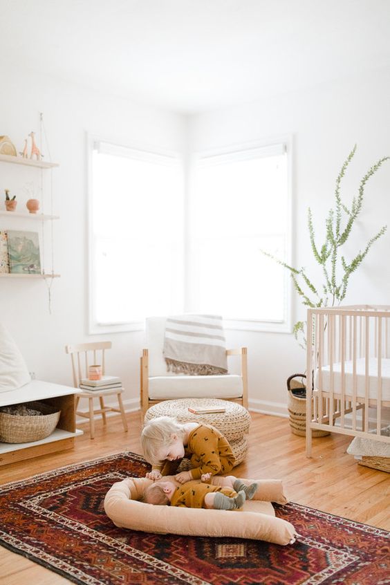 a modern boho nursery with open shelves, a stained crib, a white chair, a stool, a storage unit that doubles as a seat, a bright printed rug