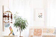 a modern boho nursery with a wodoen crib, a printed rug and bedding, a wooden pendant lamp and a stool, a white dresser, a potted plant