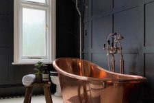 a moody black bathroom with paneling on the walls, a vintage copper bathtub and fixtures and a wooden stool and windows that bring enough light