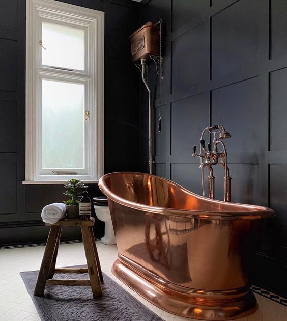 a moody black bathroom with paneling on the walls, a vintage copper bathtub and fixtures and a wooden stool and windows that bring enough light