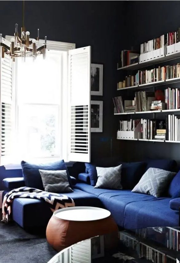 a moody living room with black walls, a navy sectional, printed pillows, open shelves, a leather coffee table, a stylsih mid-century modern chandelier
