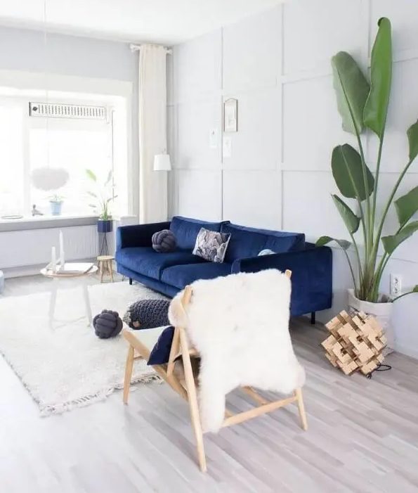 a pretty light-filled lviing room done in white and creamy, with a navy sofa, a couple of side tables, a wooden chair, a potted tree and some wood