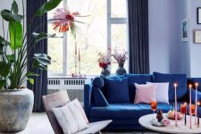 a pretty living room done with a cold color scheme, with lilac walls, a navy sofa, a round table, a lovely chair, some greenery and a sunburst chandelier