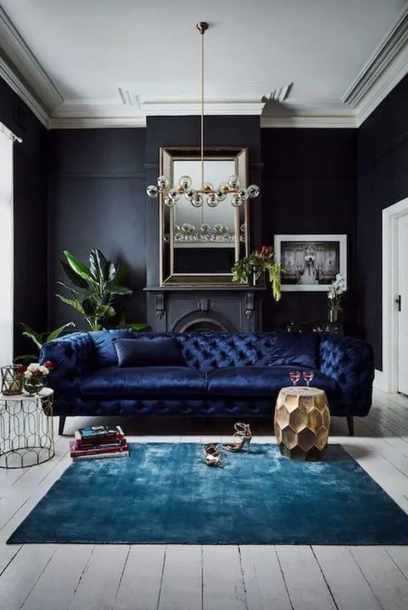 a refined living room with black walls, a non-working fireplace, a navy sofa, a bulb chandelier, a turquoise rug and a gold faceted side table and some books and plants