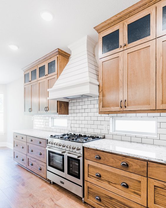 a rustic kitchen with light-stained shaker style cabinets, a white subway tile backsplash and white stone countertops, a white wood clad hood