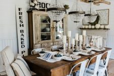 a rustic neutral dining room with a stained storage unit, a stained wooden dining table, white industrial chairs, a neutral vintage chair and a series of glam chandeliers