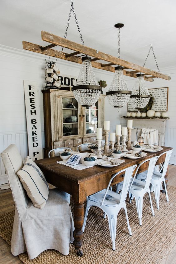 a rustic neutral dining room with a stained storage unit, a stained wooden dining table, white industrial chairs, a neutral vintage chair and a series of glam chandeliers