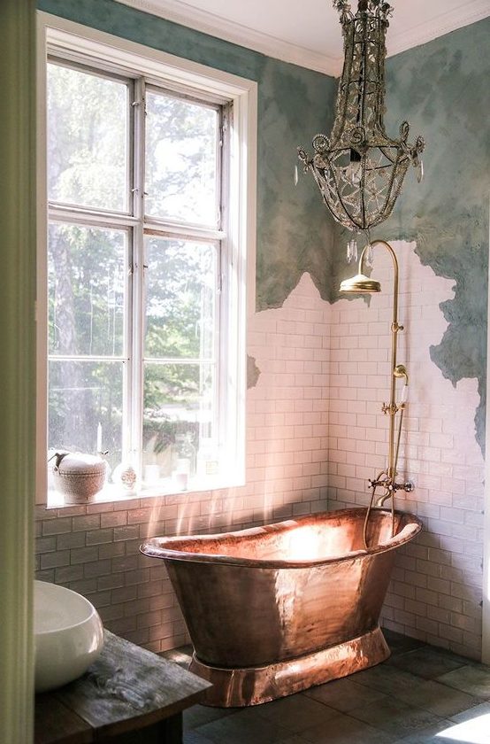 a shabby chic bathroom with green and white tile walls, a luxurious copper tub and a crystal chandelier