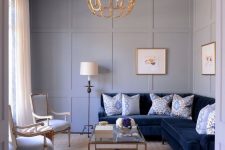 a small yet refined living room with serenity blue paneled walls, a navy sectional sofa, creamy and gold chairs, a glass coffee table and a gold chandelier