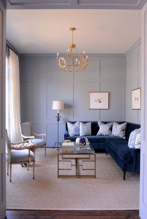 a small yet refined living room with serenity blue paneled walls, a navy sectional sofa, creamy and gold chairs, a glass coffee table and a gold chandelier
