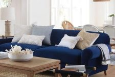 a stylish and contrasting nautical living room with a navy sofa, neutral pillows, a wooden coffee table and a folding chair, a wicker pendant lamp
