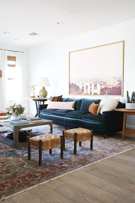 a stylish and elegant living room with a navy vlevet sofa, a statement artwork, a coffee table and woven stools, neutral pillows