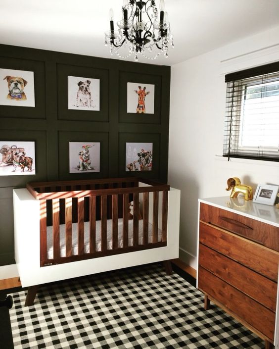 a stylish mid-century modern nursery with a black accent wall with a gallery wall, a white crib, a stained dresser, a buffalo check rug and a black chandelier