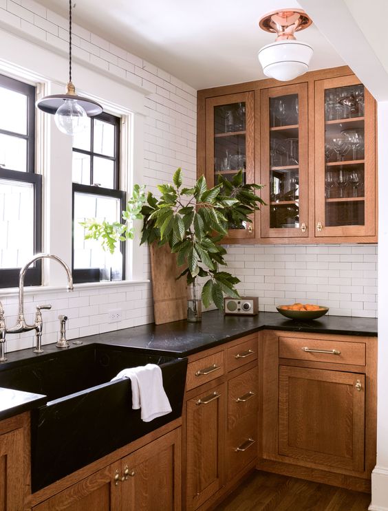 a stylish stained kitchen with black stone countertops, a white tile backsplash and neutral fixtures, greenery and glass cabinets