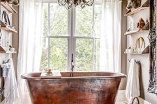 a tan bathroom with open shelves, with a large window and a copper bathtub in front of it, a chic chandelier, a rug and a basket is a gorgeous space