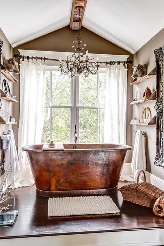 a tan bathroom with open shelves, with a large window and a copper bathtub in front of it, a chic chandelier, a rug and a basket is a gorgeous space