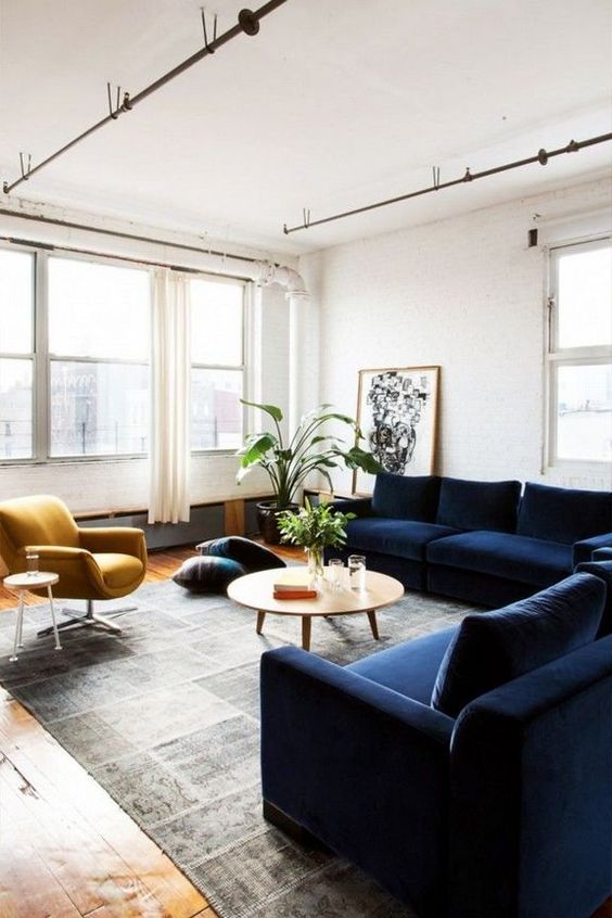 a welcoming living room with white walls, a large navy sectional, a mustard chair, a printed rug and a round table, some potted greenery