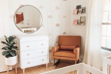 an earthy tone mid-century modern nursery with pretty wallpaper, a white dresser, an amber leather chair, a white crib, a wooden pendant lamp and books