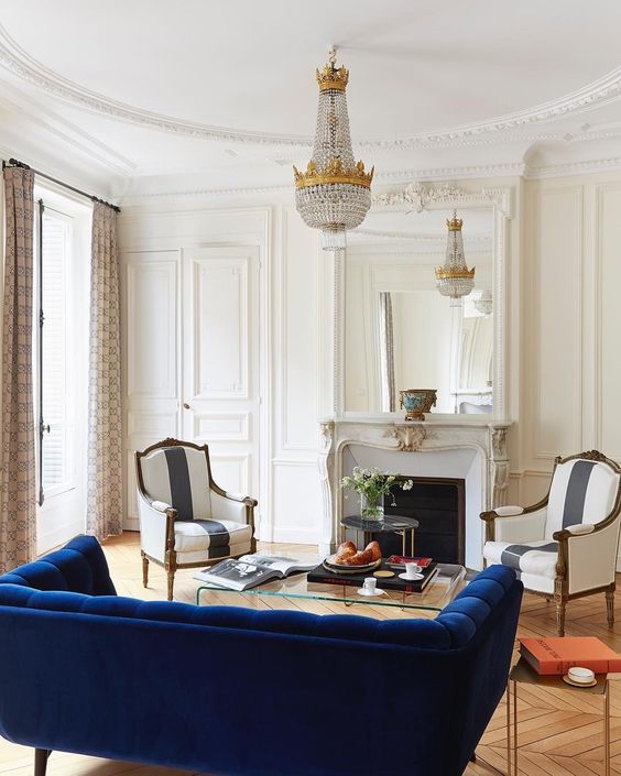 an exquisite creamy living room with a vintage fireplace, striped chairs, a heavy crystal chandelier, a navy sofa and a clear acryl coffee table