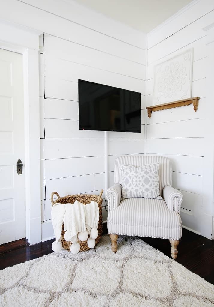 a neutral farmhouse living room with white shiplap walls, a striped vintage chair, a printed pillow, a basket with a pompom blanket, TV cords hidden in a large wire tube attached to the wall