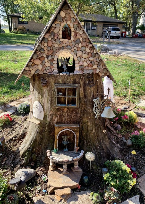 a super creative garden decoration made of a tree stump, with a pebble roof and some little elves, with blooms around