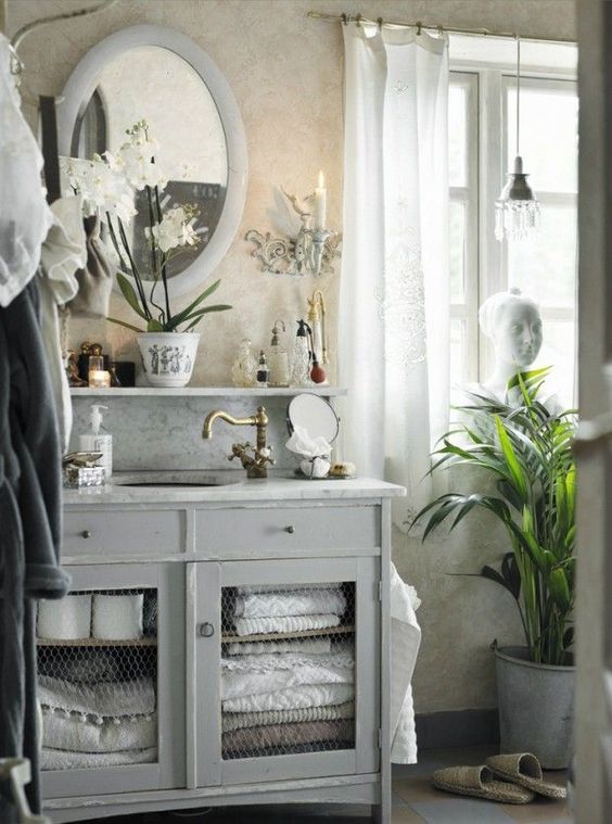 a French farmhouse bathroom with a grey vanity with storage space, an open shelf, an oval mirror in a frame and candles