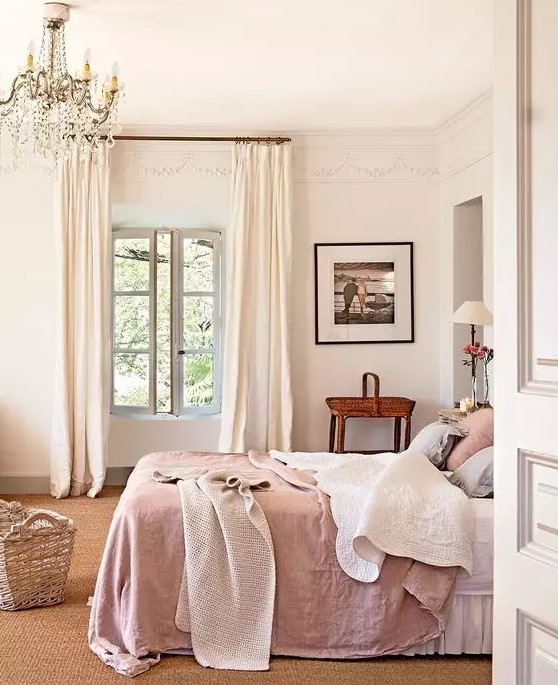 a Provence country bedroom with a bed with pink bedding, a stained table, a crystal chandelier and cool lamps and a basket for storage