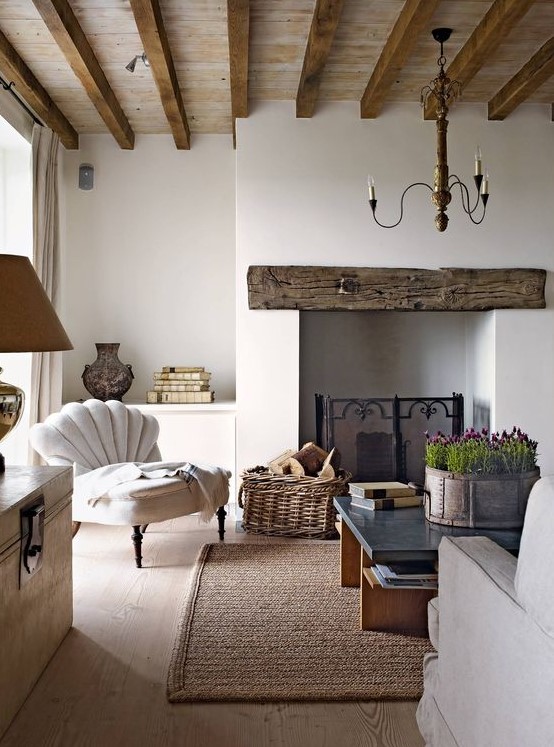 a beautiful and refined rustic living room done in neutrals, with a wooden mantel over the fireplace and wooden beams, neutral furniture, a love table and a cool chandelier