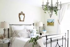 a beautiful rustic chic bedroom with a metal bed with neutral bedding, white and grey nightstands, greenery, a basket with pillows and a mirror in a chic frame