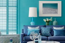 a bold beach living room with blue walls and shutters that match them, a navy sofa, printed pillows, a beach artwork, a round coffee table and a printed chair