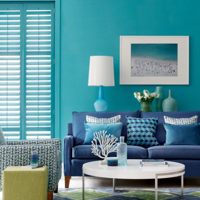 a bold beach living room with blue walls and shutters that match them, a navy sofa, printed pillows, a beach artwork, a round coffee table and a printed chair