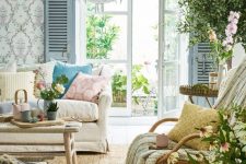 a bright Mediterranean living room with printed wallpaper, blue shutters, a neutral sofa with printed pillows, a rocker and layered rugs