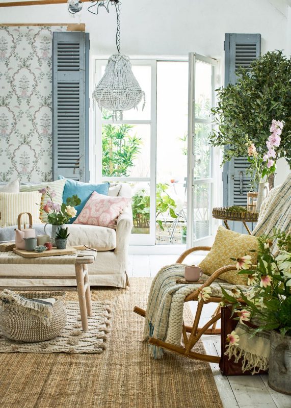 a bright Mediterranean living room with printed wallpaper, blue shutters, a neutral sofa with printed pillows, a rocker and layered rugs